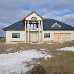 Surowy stan domu jednorodzinnego z szarą blachodachówką, jeszcze bez elewacji, z widocznymi kolumnami przy wejściu, zabezpieczonymi drewnianymi deskami oknami i błotnistym terenem wokół.