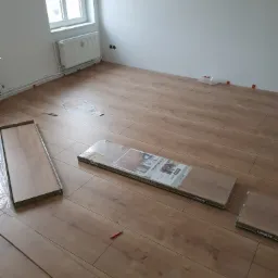 Układanie paneli podłogowych w jasnym pokoju, widoczne paczki paneli Logoclic oraz narzędzia do montażu.