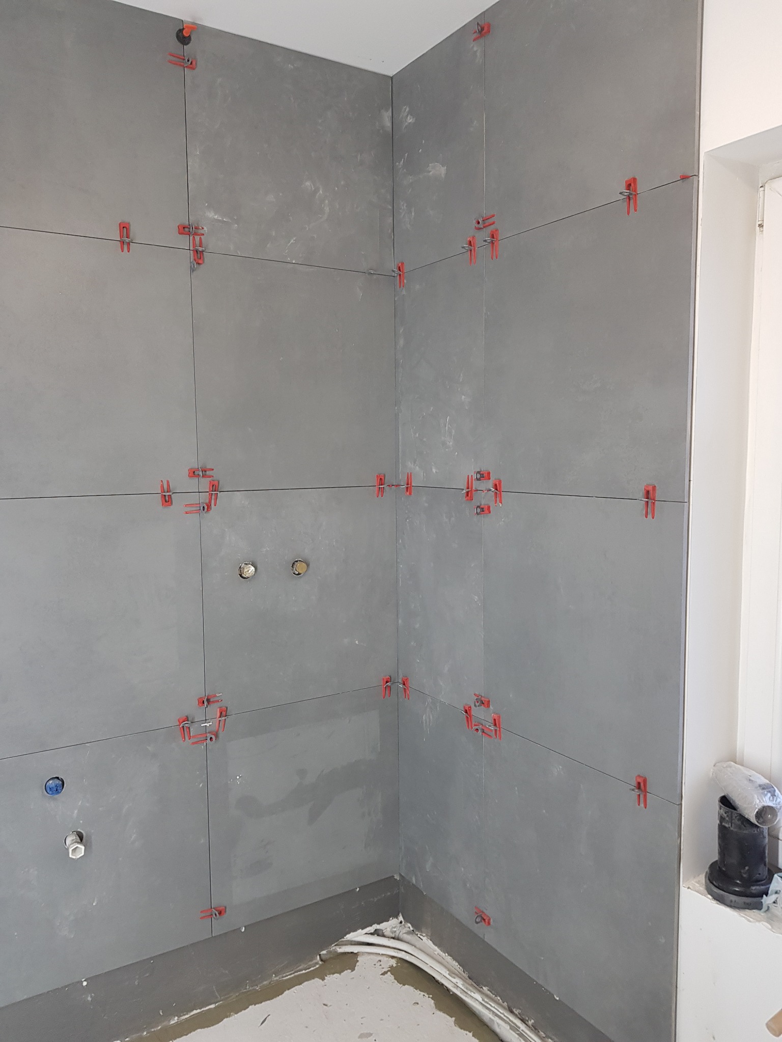 Ściana w trakcie układania szarych płytek z użyciem czerwonych klipsów poziomujących, widoczne otwory na instalacje i resztki zaprawy na podłodze.