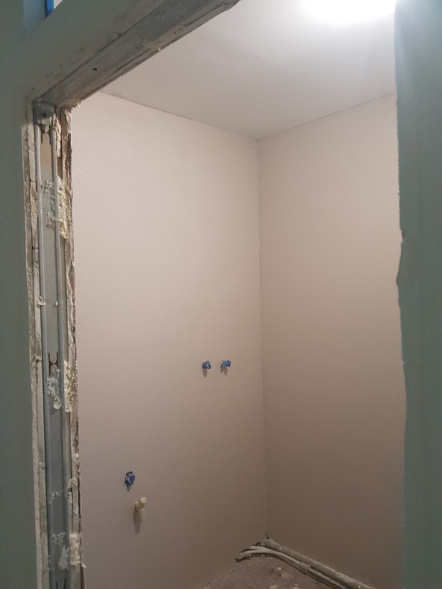 Wnętrze pomieszczenia w trakcie prac wykończeniowych, widoczne gładkie ściany w jasnym odcieniu, przygotowane do dalszych prac, z widocznymi elementami instalacji i fragmentem ościeżnicy w trakcie...