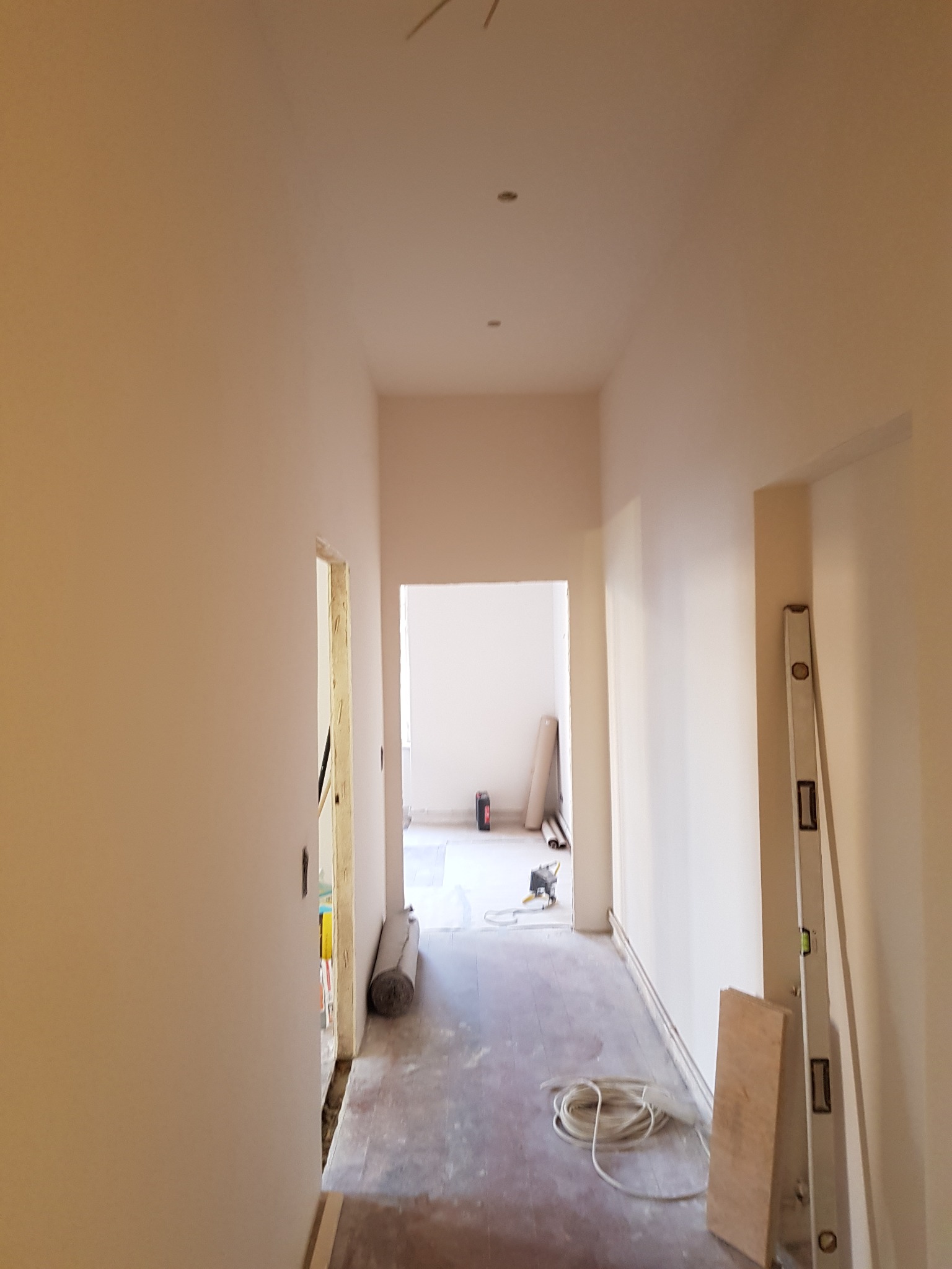 Korytarz w trakcie prac wykończeniowych, świeżo pomalowane ściany w odcieniach beżu, widoczne narzędzia i materiały budowlane, poziomica oparta o ścianę, przewody na podłodze, surowa framuga drzwi.