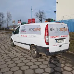 Biały samochód dostawczy firmy elektroactive z reklamą instalacji elektrycznych i odgromowych, zaparkowany na brukowanej nawierzchni, z widocznym numerem telefonu i adresem strony internetowej.