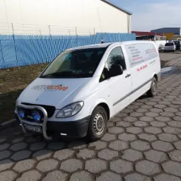 Biały samochód dostawczy z logo firmy świadczącej usługi instalacyjne, w tym odgromowe, zaparkowany na kostce brukowej przed budynkiem przemysłowym i niebieskim płotem, z dodatkowymi lampami...