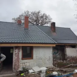 Dom w trakcie remontu elewacji i wymiany dachu, widoczne nowe okno i gruz na podwórku.