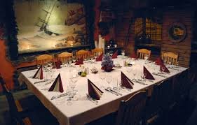 Długi stół nakryty białym obrusem, ustawiony dla kilkunastu osób, z bordowymi serwetkami, kieliszkami do wina i dekoracjami świątecznymi w tle ciemnego, klimatycznego wnętrza restauracji z obrazem...