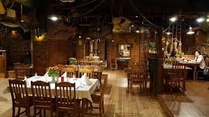 Wnętrze restauracji w rustykalnym stylu z drewnianymi meblami i dekoracjami, nakryte stoły z białymi obrusami i kwiatami, widoczni goście przy stoliku w tle.