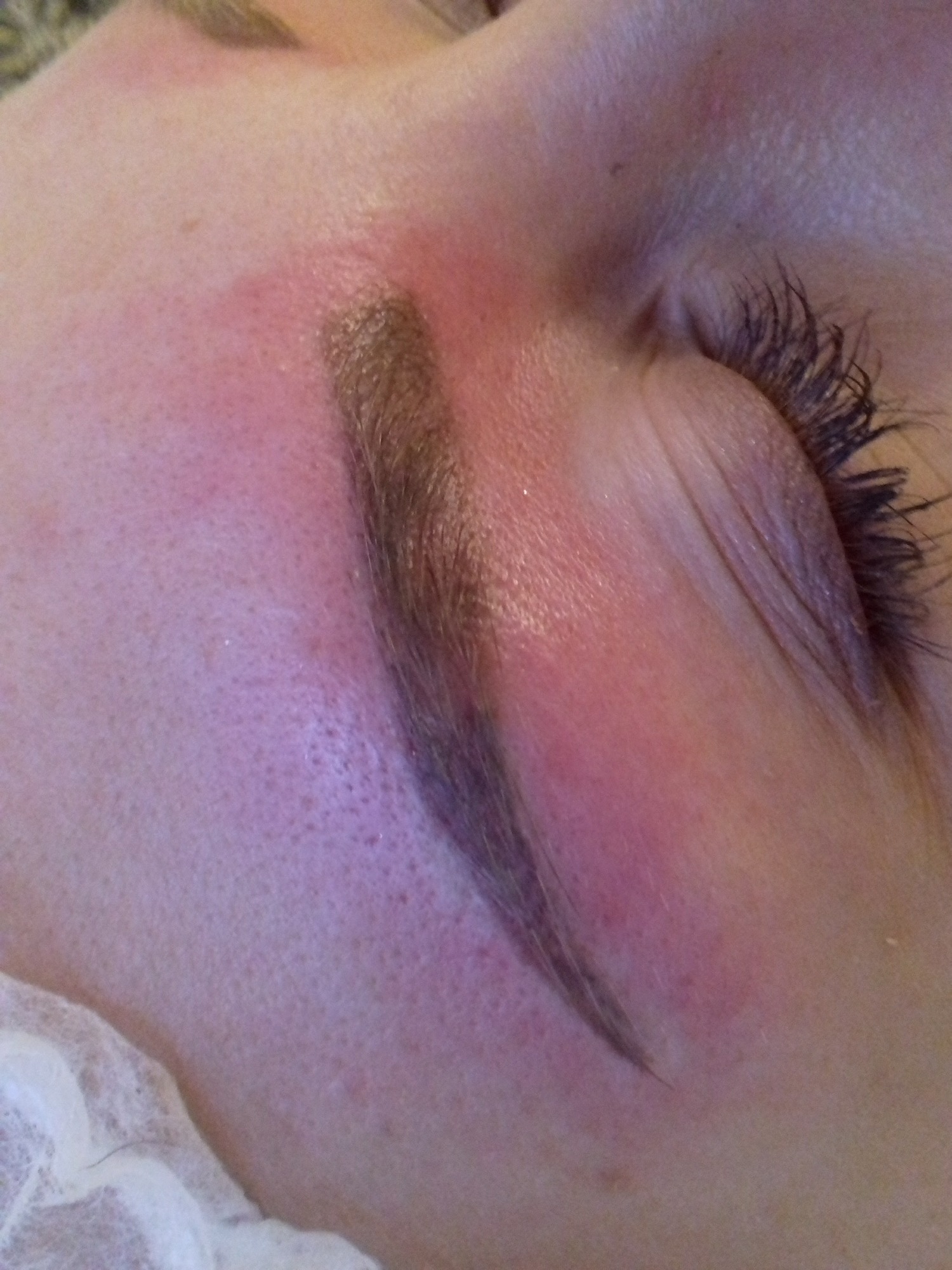 Zbliżenie na oko i brwi po zabiegu pigmentacji, widoczna zaczerwieniona skóra wokół brwi, pojedyncze rzęsy i fragment jednorazowego podkładu pod głowę.