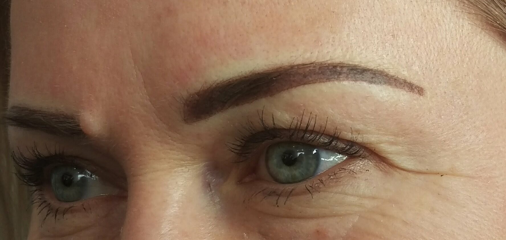Zbliżenie na oko kobiety z widocznym makijażem permanentnym brwi; widoczne zmarszczki mimiczne i lekko błyszcząca skóra.