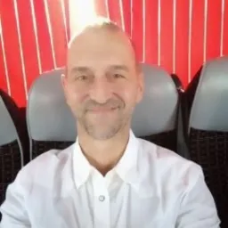 Portret mężczyzny w białej koszuli na tle czerwonych zasłon i szarych foteli, oświetlonego od góry.