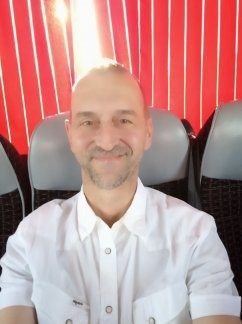 Portret mężczyzny w białej koszuli na tle czerwonych zasłon i szarych foteli, oświetlonego od góry.