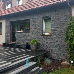 Tomasz - Elewacja domu wykończona szarą kamienną okładziną, z drewnianym tarasem, roślinami w donicach i butelką wody na schodach.