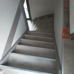 Kątowe schody wykończone panelami imitującymi drewno, z szarymi listwami przypodłogowymi i fragmentem ściany z cegły w tle.