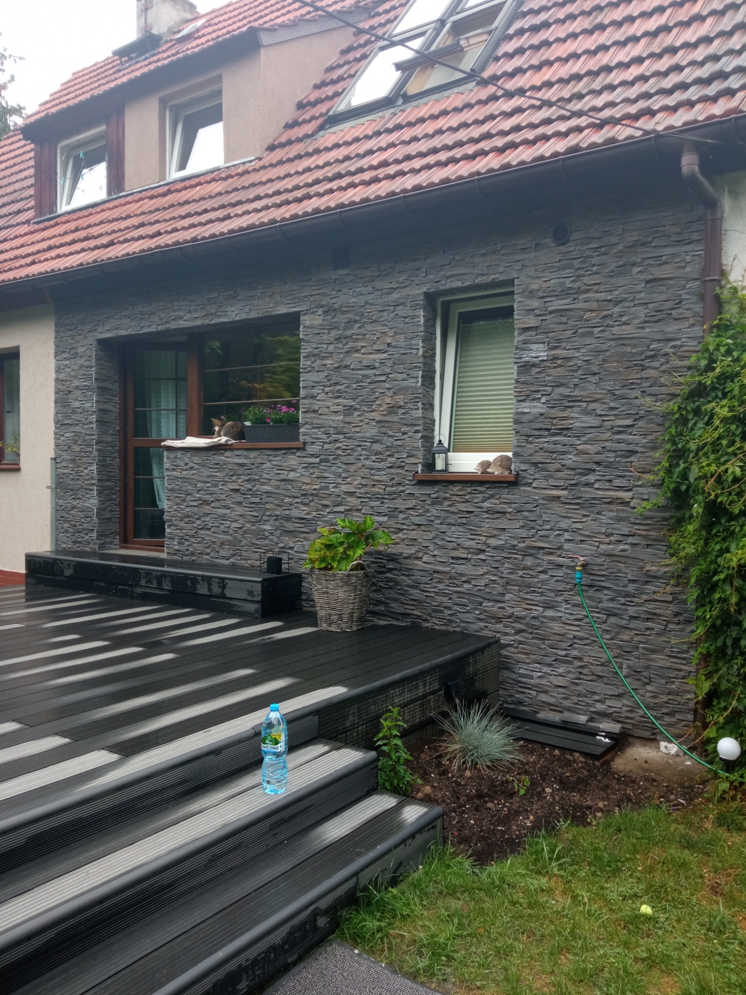 Elewacja domu wykończona szarą kamienną okładziną, z drewnianym tarasem, roślinami w donicach i butelką wody na schodach.