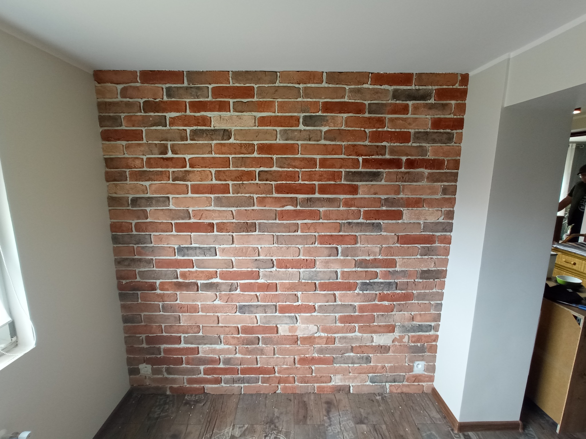 Ściana z dekoracyjnej cegły w odcieniach czerwieni i brązu, na podłodze panele imitujące drewno, widoczny fragment gniazdka elektrycznego przy podłodze.