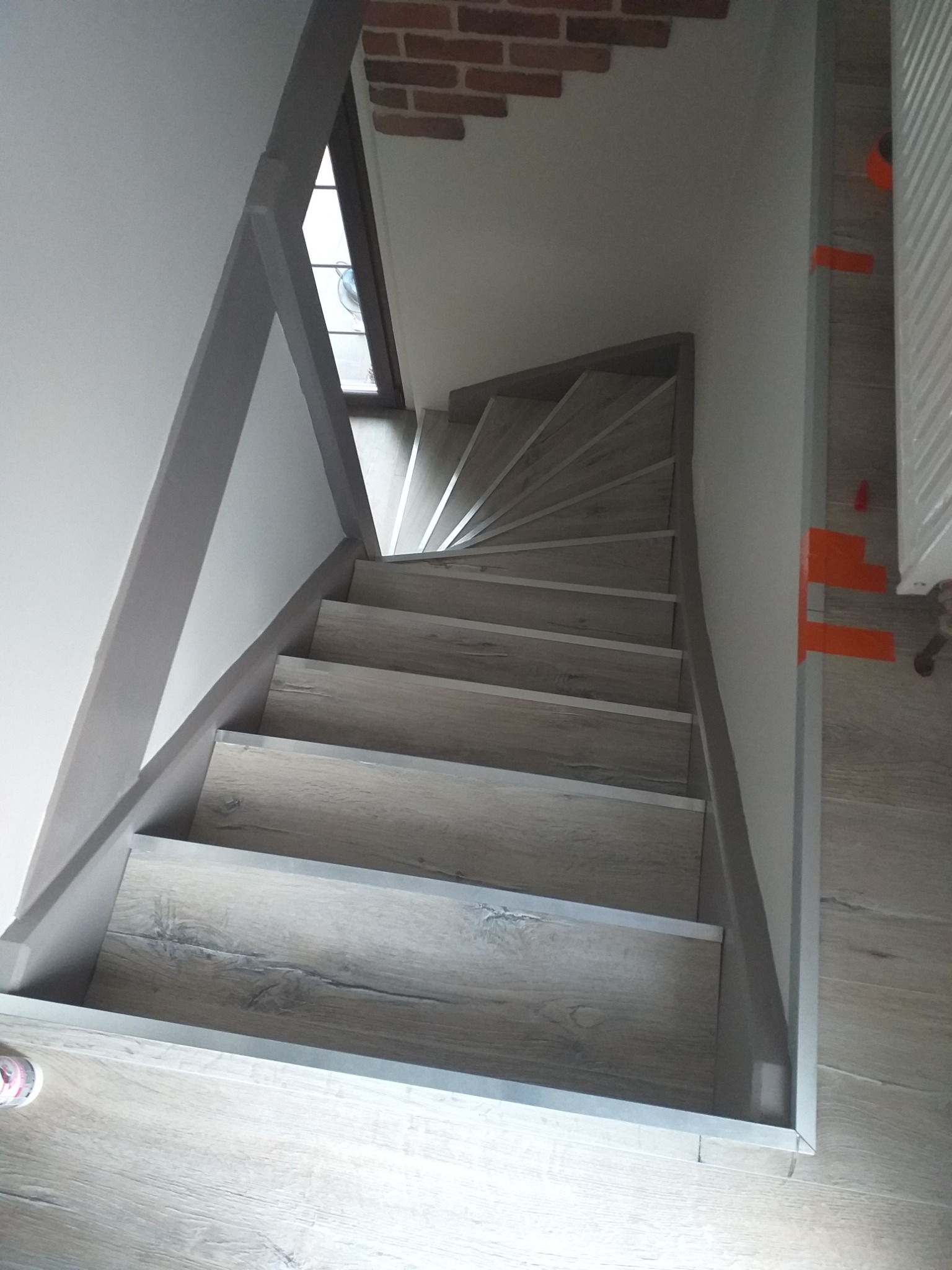 Kątowe schody wykończone panelami imitującymi drewno, z szarymi listwami przypodłogowymi i fragmentem ściany z cegły w tle.