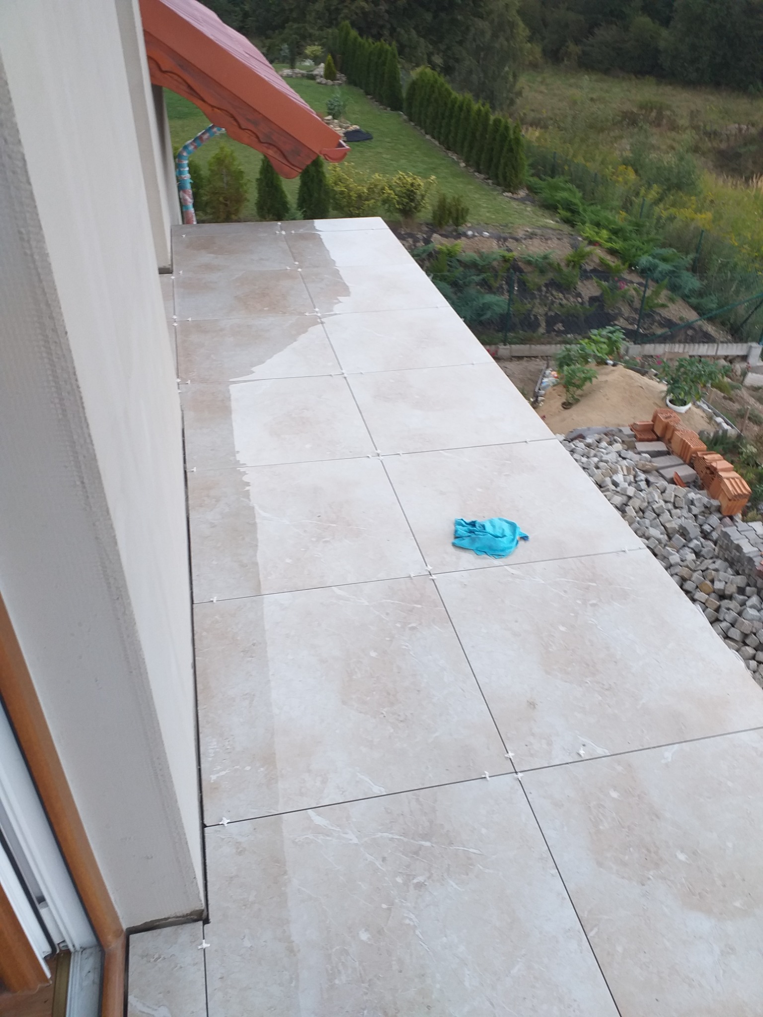 Jasne płytki gresowe na balkonie w trakcie układania, widoczne krzyżyki dystansowe, niebieska ścierka i materiały budowlane, z widokiem na ogród za balustradą.