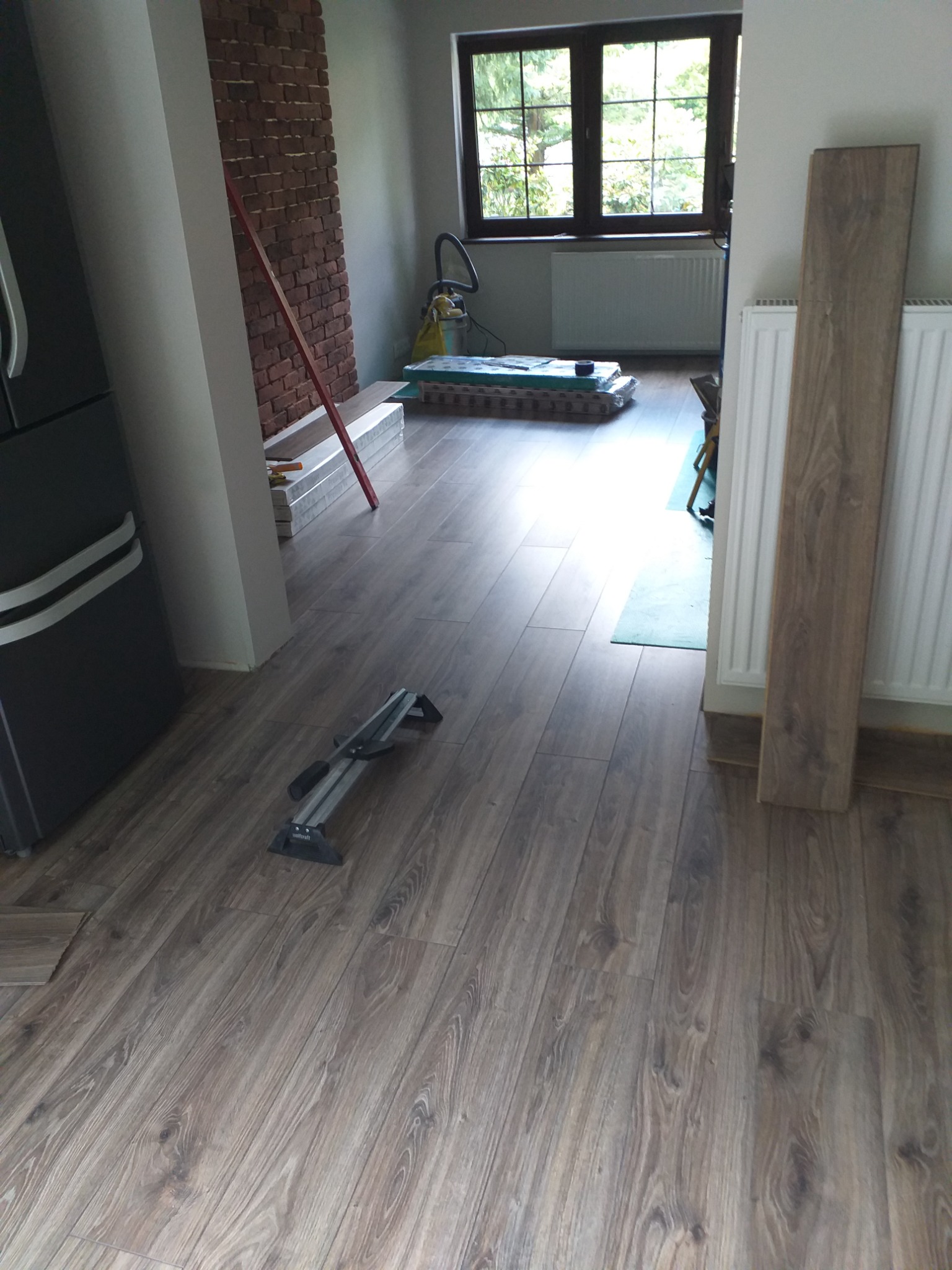 Fragment pokoju z ułożonymi panelami podłogowymi o wzorze drewna, obok ściany z cegły i narzędzia do cięcia paneli na podłodze.