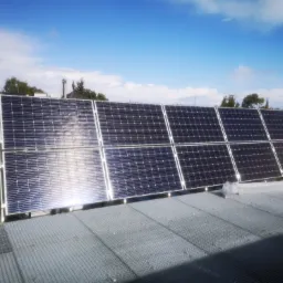 Rząd paneli słonecznych na dachu budynku, widoczny z perspektywy lekko od dołu, z metalową kratą pod spodem i budynkami w tle pod błękitnym niebem.