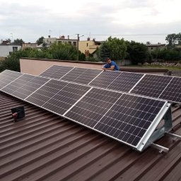 Mono Energy Sp. z o.o. - Panele słoneczne zamontowane na brązowym dachu z blachy trapezowej, instalator w niebieskiej koszulce opiera się o konstrukcję, w tle zabudowa jednorodzinna i pochmurne niebo.