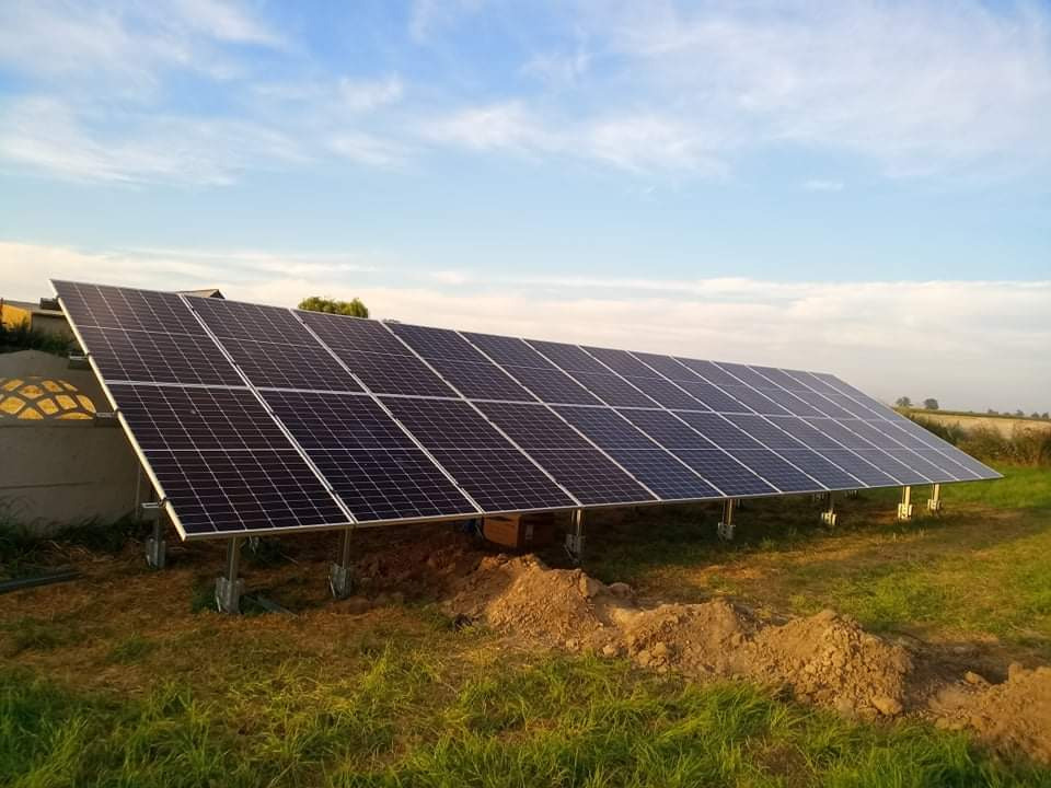 Rząd paneli słonecznych na metalowej konstrukcji, widoczny na tle pola i błękitnego nieba, z wykopaną ziemią u podstawy konstrukcji.