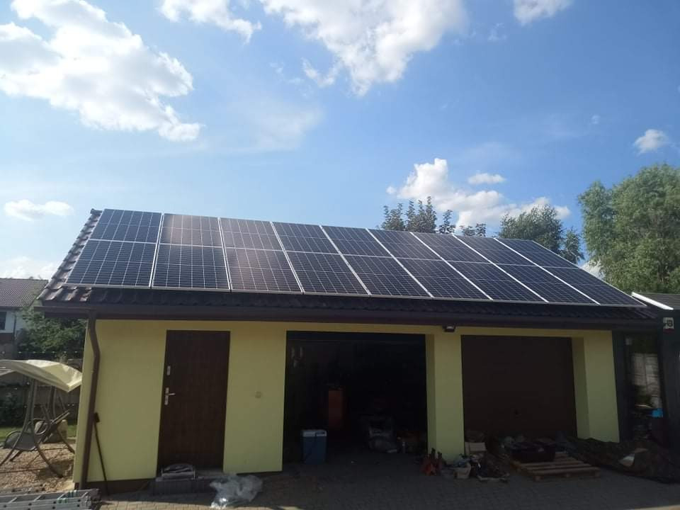 Żółty garaż z brązową bramą i panelami słonecznymi na dachu, widok z dołu na tle błękitnego nieba z chmurami.