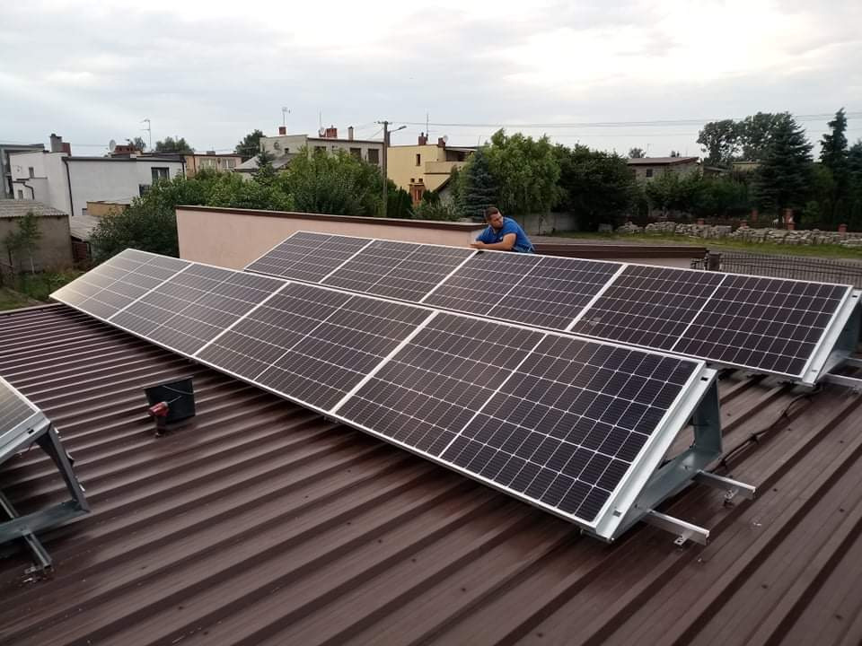 Panele słoneczne zamontowane na brązowym dachu z blachy trapezowej, instalator w niebieskiej koszulce opiera się o konstrukcję, w tle zabudowa jednorodzinna i pochmurne niebo.
