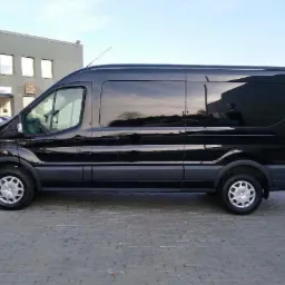 Czarny, długi bus Ford Transit z przyciemnionymi szybami, zaparkowany na kostce brukowej przed salonem samochodowym. W oknach odbija się budynek i drzewa.