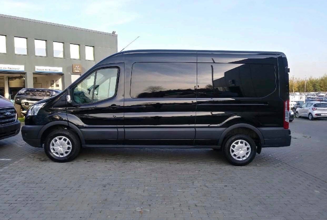 Czarny, długi bus Ford Transit z przyciemnionymi szybami, zaparkowany na kostce brukowej przed salonem samochodowym. W oknach odbija się budynek i drzewa.