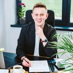 Uśmiechnięty mężczyzna w marynarce siedzi za biurkiem, podpisuje dokument, obok laptop, kawa i roślina doniczkowa.