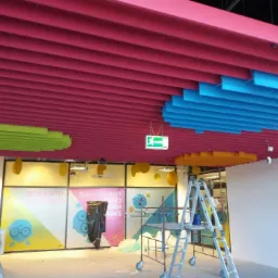 Kolorowy sufit rastrowy z elementami w odcieniach różu, niebieskiego, zielonego i żółtego, widoczne rusztowanie i elementy wykończeniowe.