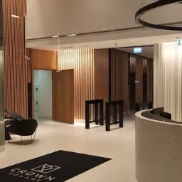 Wnętrze biurowca z drewnianymi panelami ściennymi, widoczne drzwi, barierki wejściowe i okrągłe lampy sufitowe, na podłodze wycieraczka z logo Crown Square.