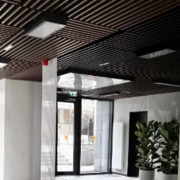 Nowoczesny hol wejściowy z drewnianym sufitem lamelowym, białymi ścianami z delikatną fakturą, marmurową podłogą, szklanymi drzwiami z pomarańczowymi oznaczeniami i roślinami doniczkowymi.