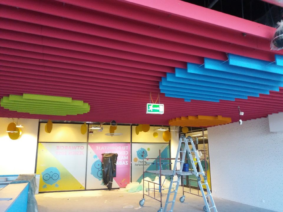 Kolorowy sufit rastrowy z elementami w odcieniach różu, niebieskiego, zielonego i żółtego, widoczne rusztowanie i elementy wykończeniowe.