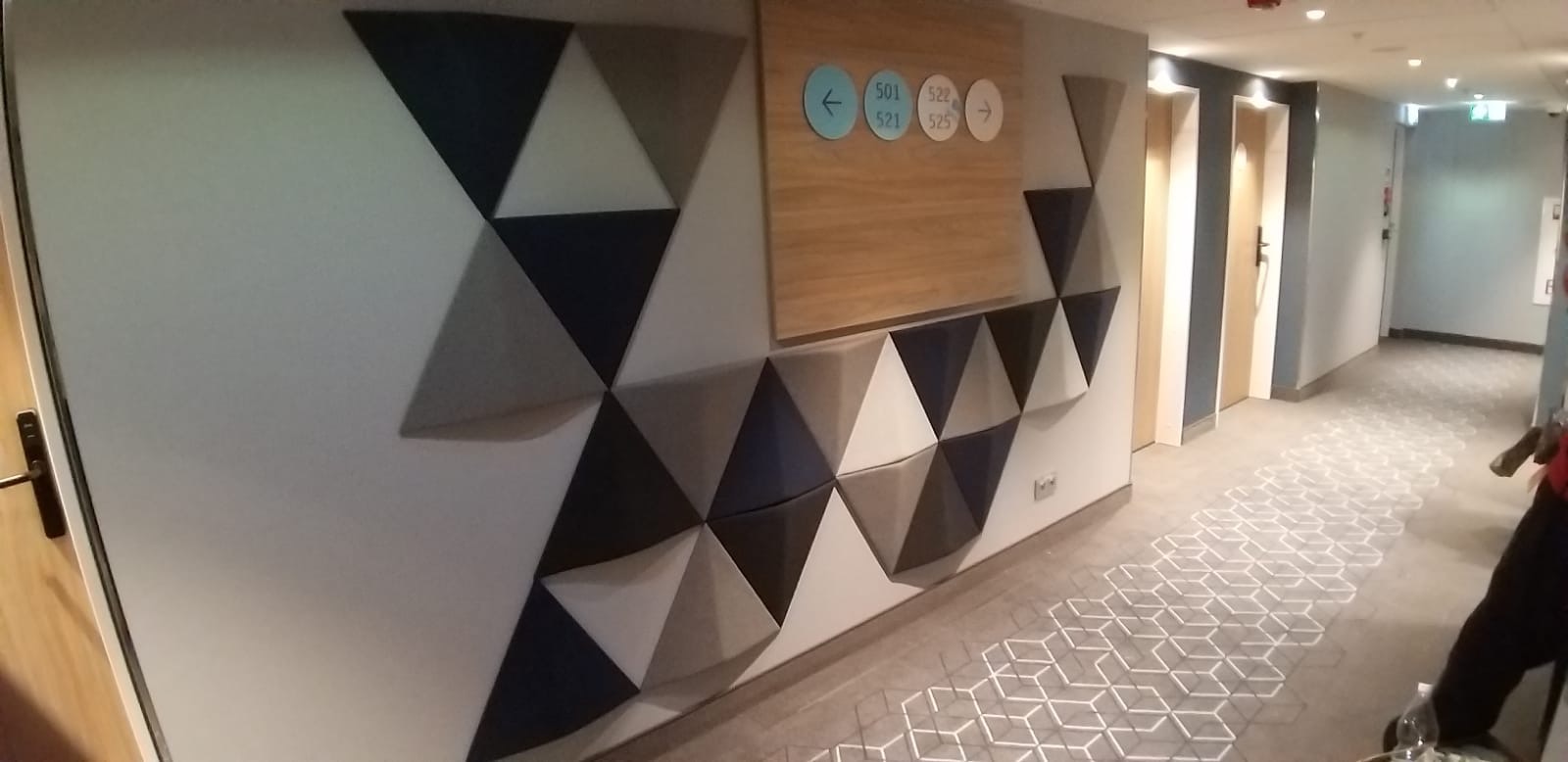 Korytarz hotelowy z panelami akustycznymi w kształcie trójkątów w odcieniach szarości i niebieskiego na ścianie, drewnianą tabliczką informacyjną z numerami pokoi i wzorzystą wykładziną podłogową.