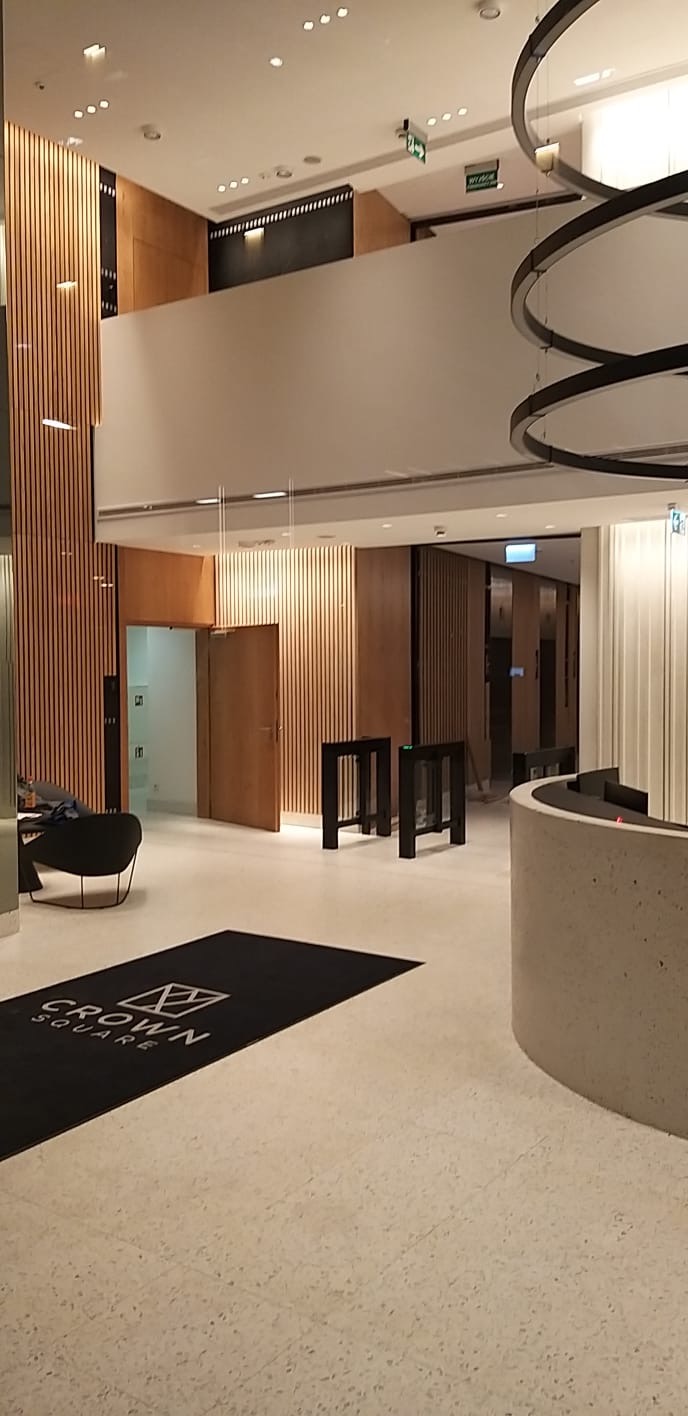 Wnętrze biurowca z drewnianymi panelami ściennymi, widoczne drzwi, barierki wejściowe i okrągłe lampy sufitowe, na podłodze wycieraczka z logo Crown Square.