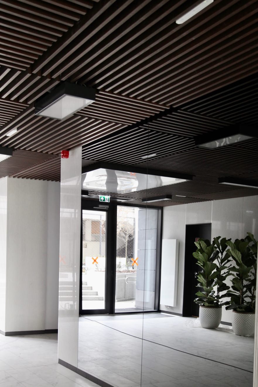 Nowoczesny hol wejściowy z drewnianym sufitem lamelowym, białymi ścianami z delikatną fakturą, marmurową podłogą, szklanymi drzwiami z pomarańczowymi oznaczeniami i roślinami doniczkowymi.