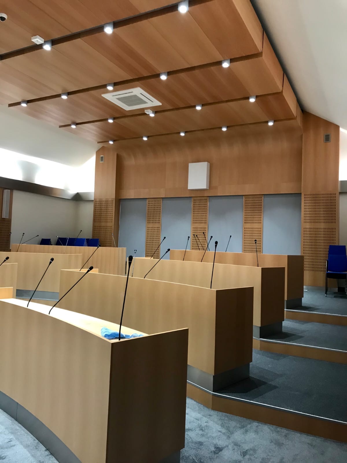 Sala konferencyjna z drewnianym wykończeniem ścian i sufitu, rzędy jasnych mównic z mikrofonami, szara wykładzina podłogowa i nowoczesne oświetlenie.