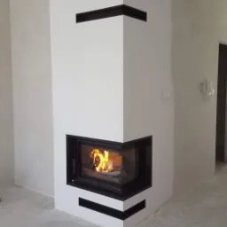 Nowoczesny narożny wkład kominkowy z czarną obudową i płomieniami, umieszczony w białej, minimalistycznej zabudowie z poziomymi szczelinami wentylacyjnymi.