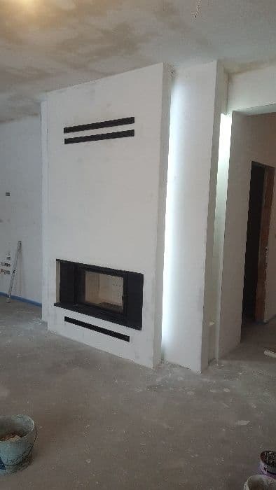 Nowoczesna obudowa wkładu kominkowego w trakcie wykończenia, surowy beton na podłodze, widoczne ślady prac budowlanych, minimalistyczny design z czarnymi akcentami.