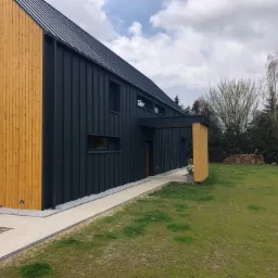 Nowoczesny dom w Czeszowie z elewacją w kolorze grafitu i drewna, otoczony trawnikiem i ścieżką z jasnej kostki. Osoba w kamuflażu klęczy przy kostce. W tle widoczne drzewa.