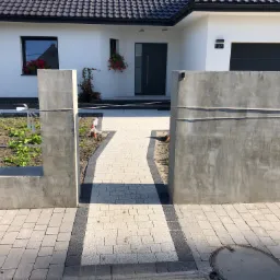 Nowoczesny podjazd z jasnoszarej kostki brukowej z ciemnoszarymi obrzeżami, prowadzący do białego domu z szarą stolarką i numerem 12A, otoczony surowymi, betonowymi elementami ogrodzenia.