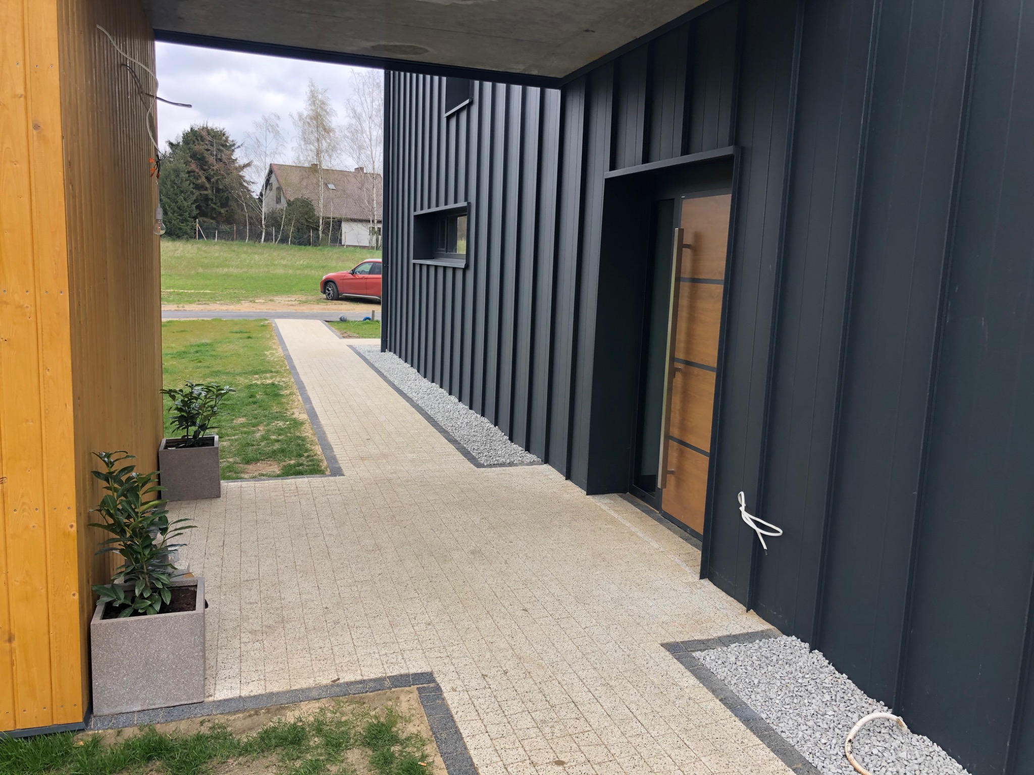 Nowoczesna posesja z jasną kostką brukową, ciemną elewacją z pionowych paneli i minimalistycznym ogrodem z roślinami w donicach.