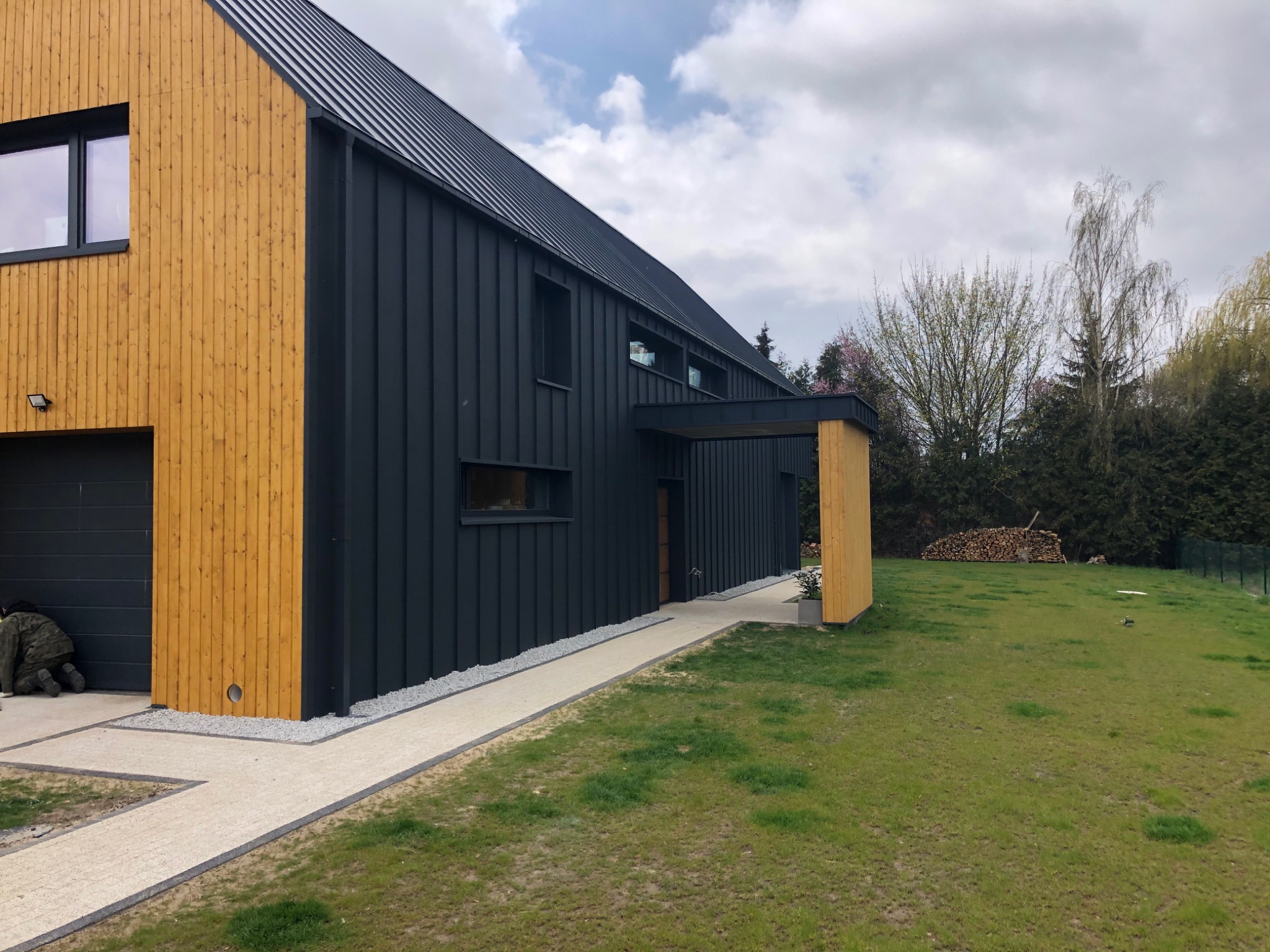 Nowoczesny dom w Czeszowie z elewacją w kolorze grafitu i drewna, otoczony trawnikiem i ścieżką z jasnej kostki. Osoba w kamuflażu klęczy przy kostce. W tle widoczne drzewa.