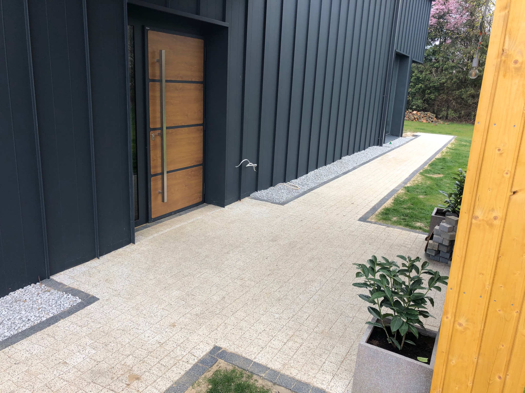 Jasna kostka brukowa prowadząca do nowoczesnego domu z ciemną elewacją, obrzeża z szarego grysu, donice z roślinami.