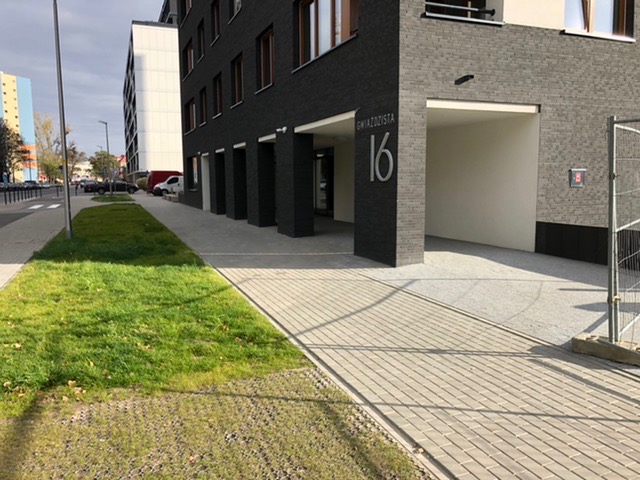 Chodnik z szarej kostki brukowej prowadzący do budynku z ciemnej cegły, trawnik z kratką trawnikową po lewej stronie, adres Gwiaździsta 16 na filarze.