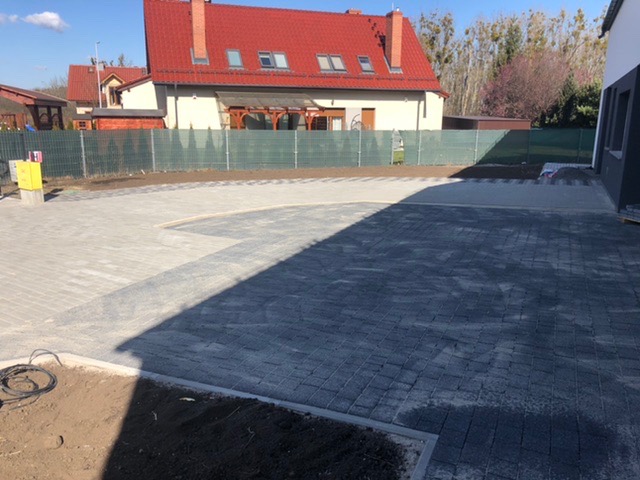 Ułożona kostka brukowa w dwóch odcieniach szarości tworząca podjazd przed domem z czerwonym dachem, widoczny kabel i fragment rabaty.