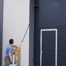 Mężczyzna czyści ciemną, poziomą bramę garażową za pomocą szczotki na długim wysięgniku i mobilnego systemu filtracji wody Ionic Trion; mokra kostka brukowa przed budynkiem.
