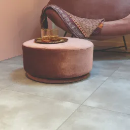 Jasnoszare płytki podłogowe z widocznym szwem, uzupełnione różowym pufem z dwoma szklankami na złotej tacy oraz fragmentem różowego fotela z narzutą w tle.