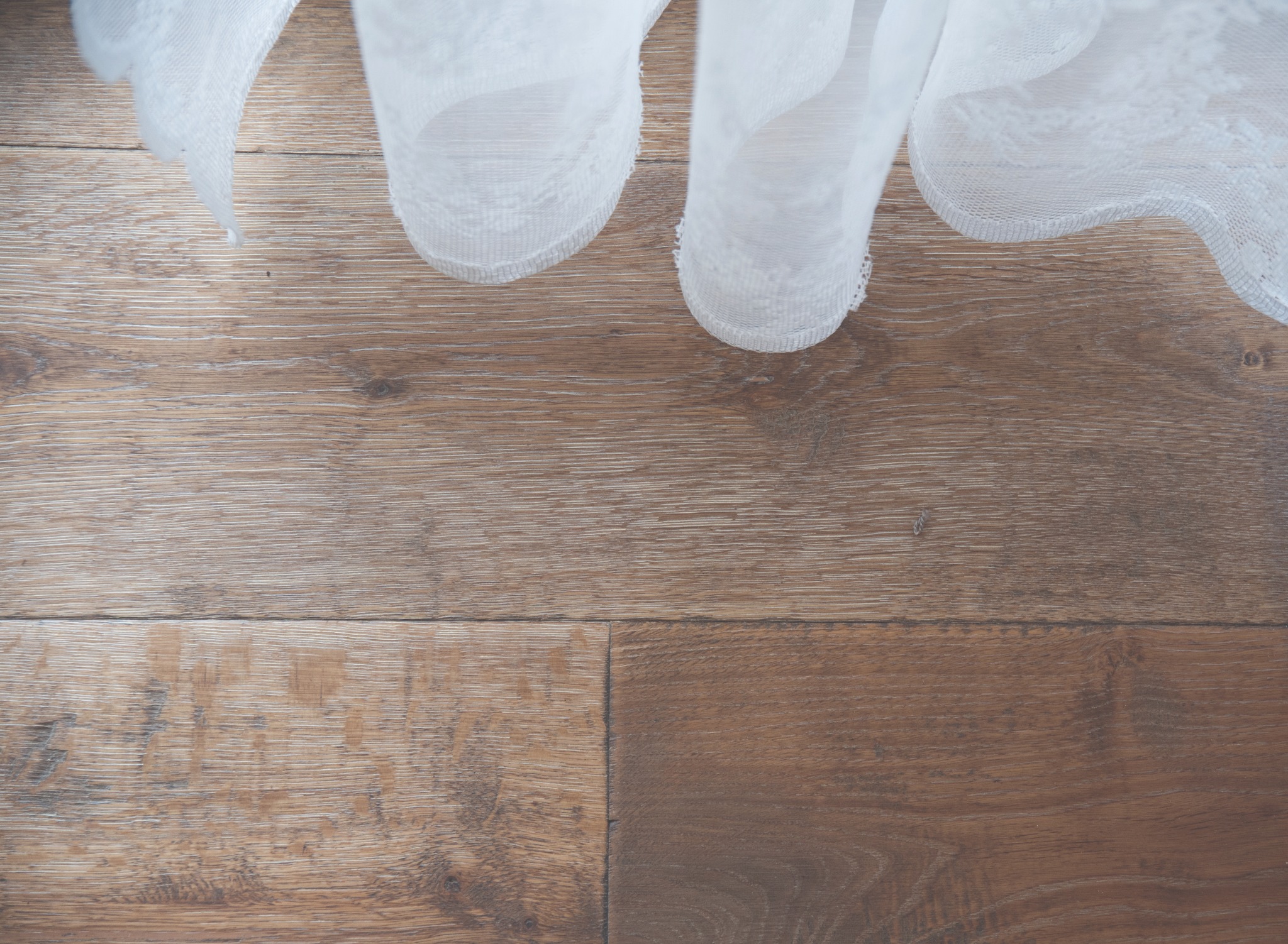 Jasnobrązowa podłoga z paneli drewnianych z delikatną teksturą, częściowo przykryta zwiewną, białą firanką z koronkowym wykończeniem w górnej części kadru.