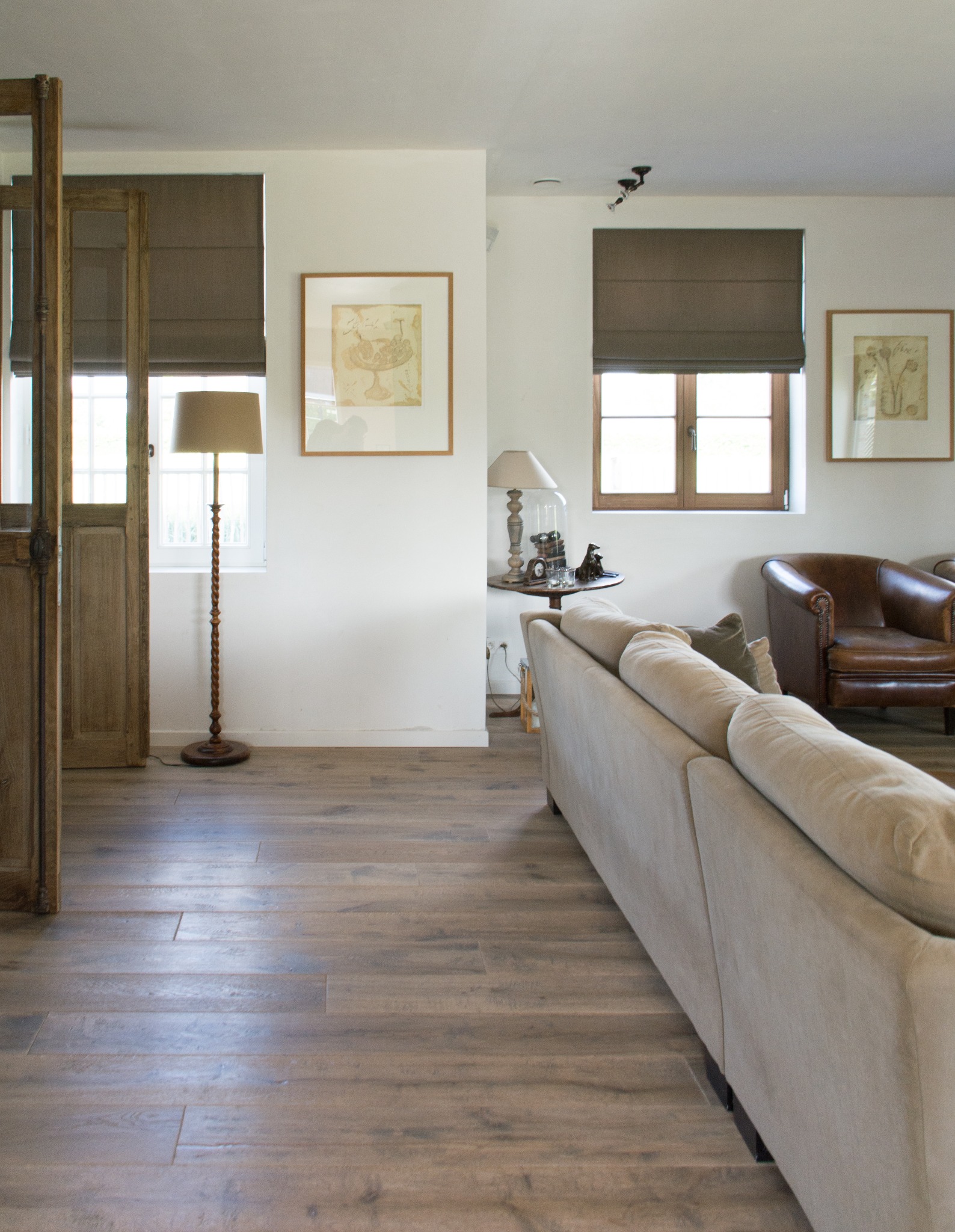 Jasne wnętrze salonu z drewnianą podłogą ułożoną w deski, widoczne drzwi z przeszklonymi szybami, sofa, skórzany fotel i lampa stojąca.